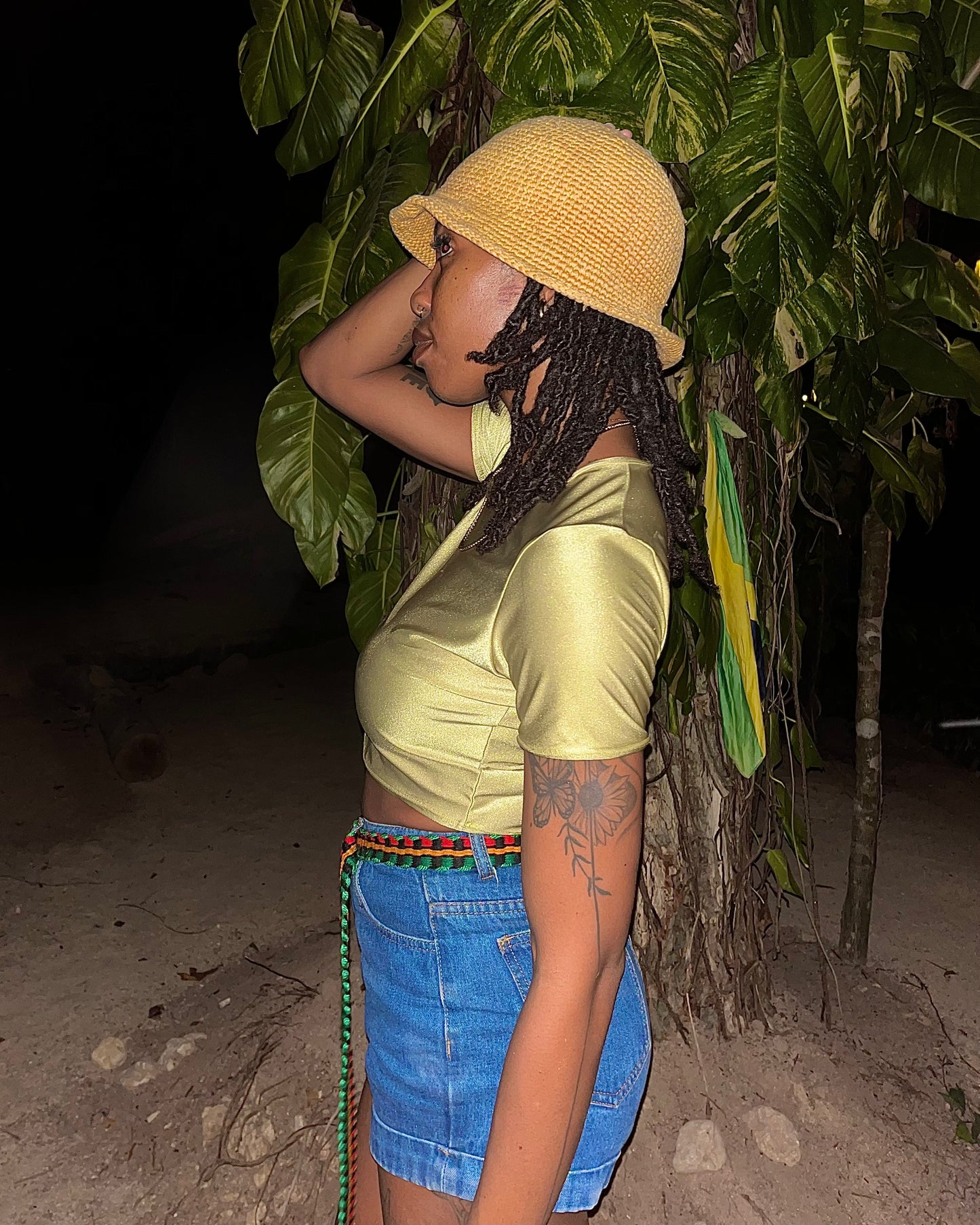 A woman in a yellow bucket hat, shiny pale yellow crop top, and blue denim shorts stands outdoors at night beside a palm tree, with a green, yellow, and black flag hanging from the tree trunk.
