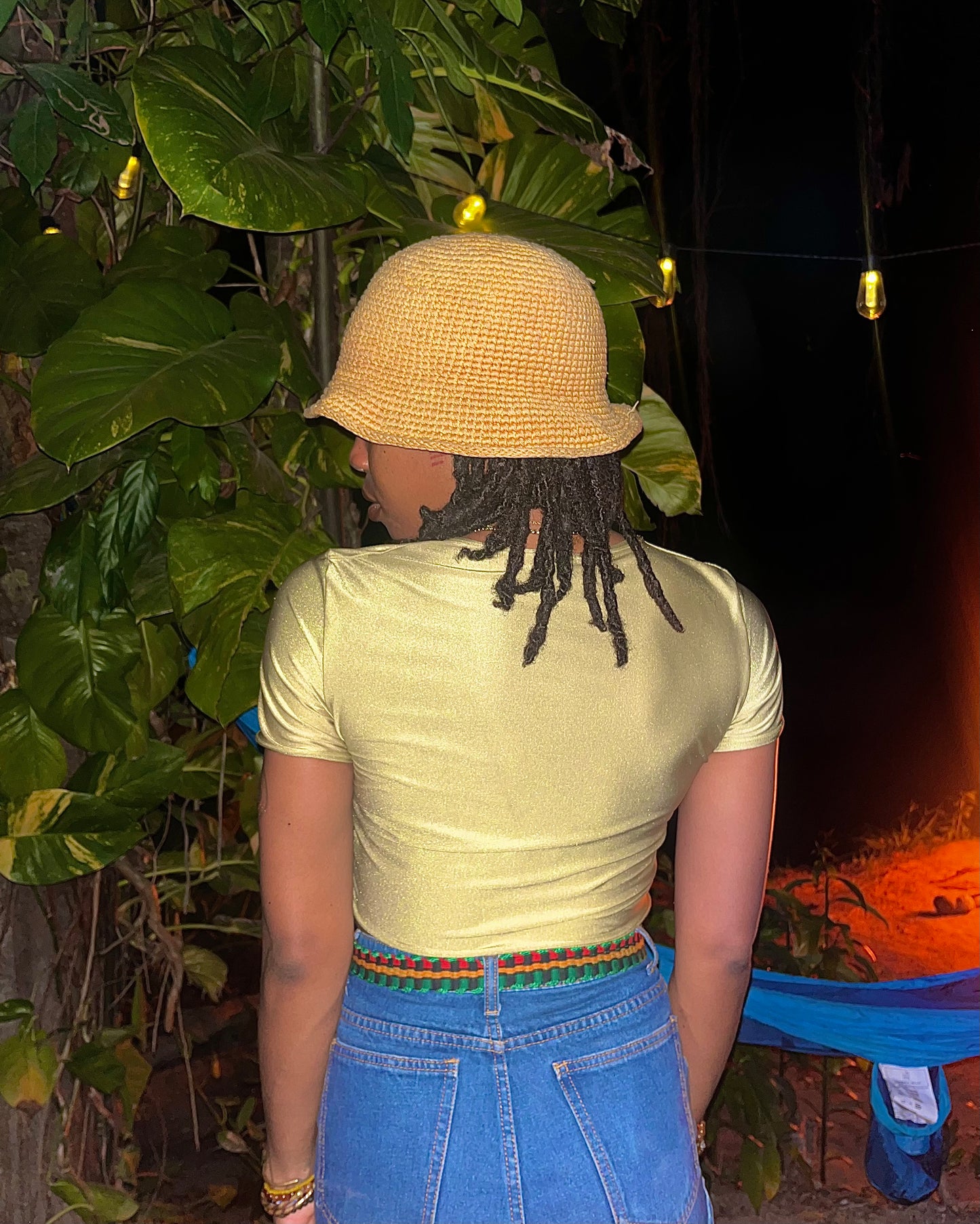 A person viewed from behind wearing a light yellow short-sleeve crop top, a beige woven bucket hat, and blue jeans with a colorful braided belt, standing outdoors at night among large green leaves and warm string lights.