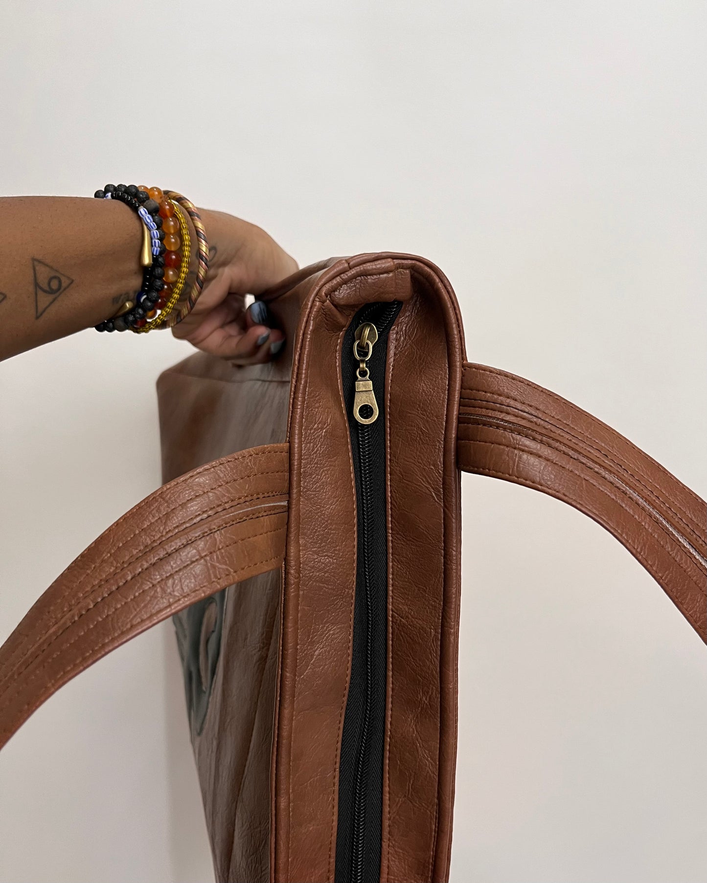 A close-up of a brown faux-leather tote bag with a black zipper and gold pull, held by a person's hand adorned with colorful beaded bracelets against a plain off-white background.
