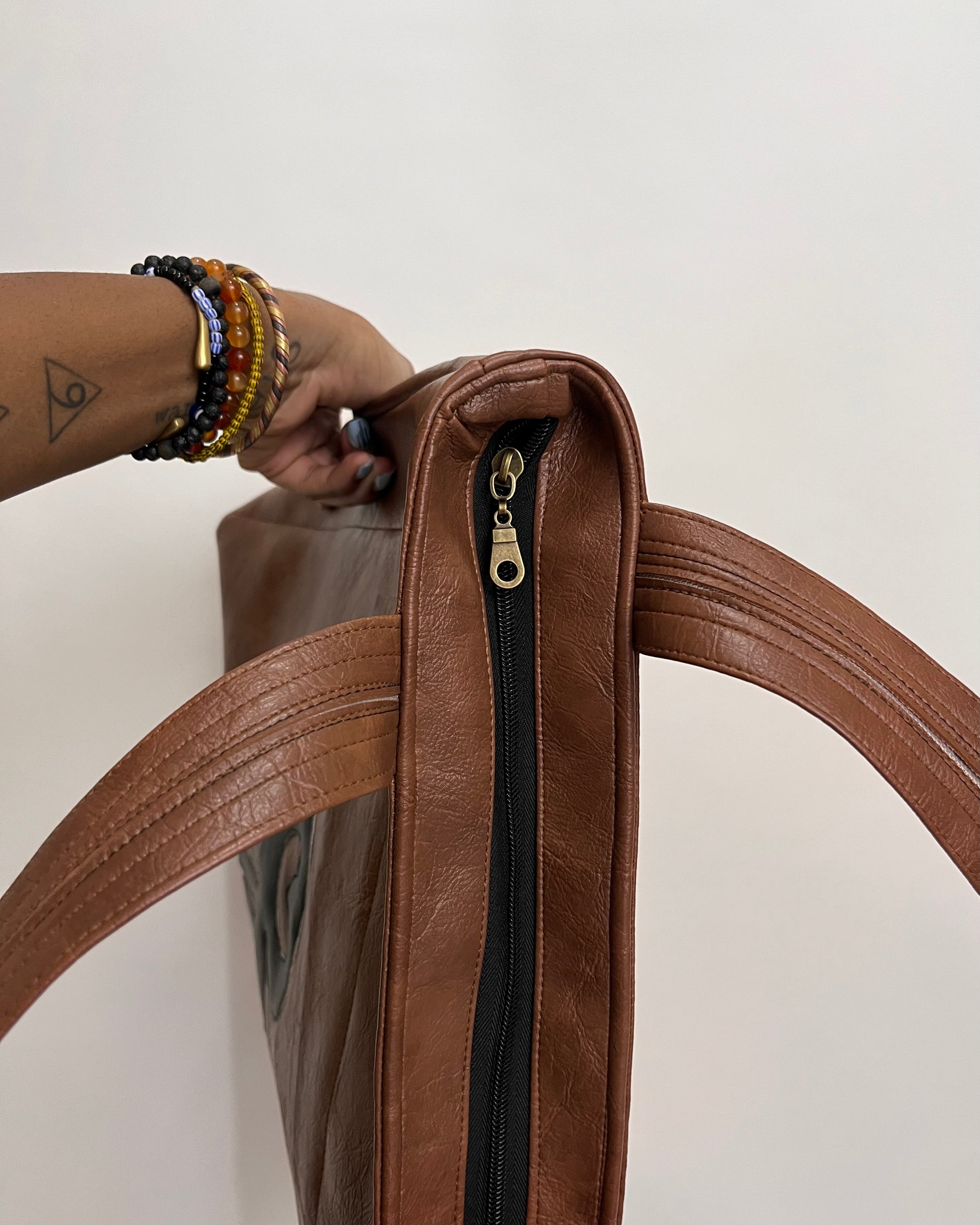 A close-up of a brown faux-leather tote bag with a black zipper and gold pull, held by a person's hand adorned with colorful beaded bracelets against a plain off-white background.