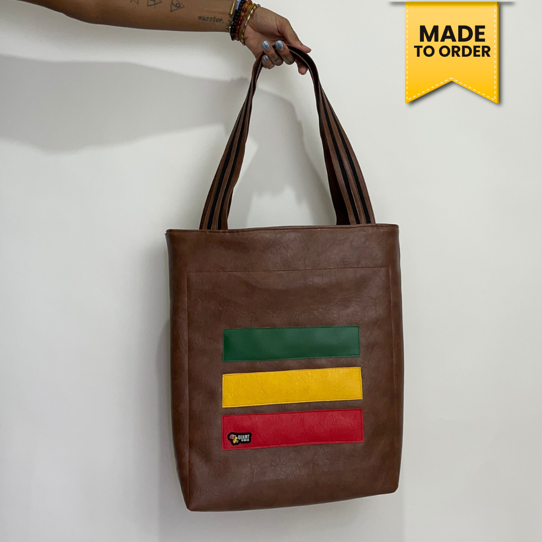 A brown faux-leather tote bag with a green, yellow, and red striped graphic on the front, held by a hand against a white wall. A yellow 'MADE TO ORDER' badge is in the top right corner.