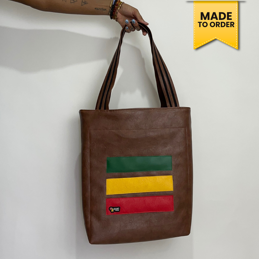 A brown faux-leather tote bag with a green, yellow, and red striped graphic on the front, held by a hand against a white wall. A yellow 'MADE TO ORDER' badge is in the top right corner.